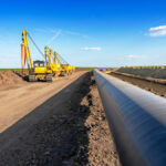 A silver pipeline runs through a newly dug dirt path with green fields on both sides. Yellow excavators are next to the pipes.