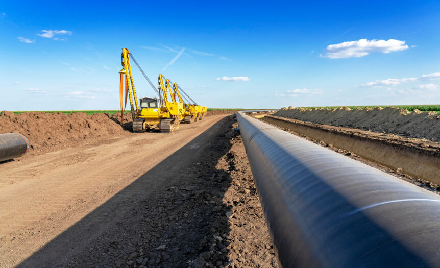 A silver pipeline runs through a newly dug dirt path with green fields on both sides. Yellow excavators are next to the pipes.