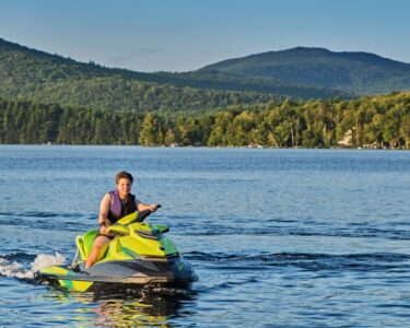 A person in a purple life jacket driving a green jet ski on a body of water surrounded by a dense forest.