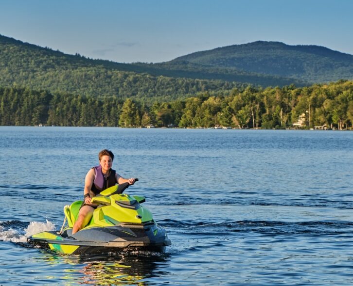 A person in a purple life jacket driving a green jet ski on a body of water surrounded by a dense forest.