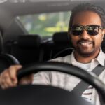 A man wearing a white shirt and black sunglasses sits in the driver's seat of a vehicle. His hands are on the steering wheel.