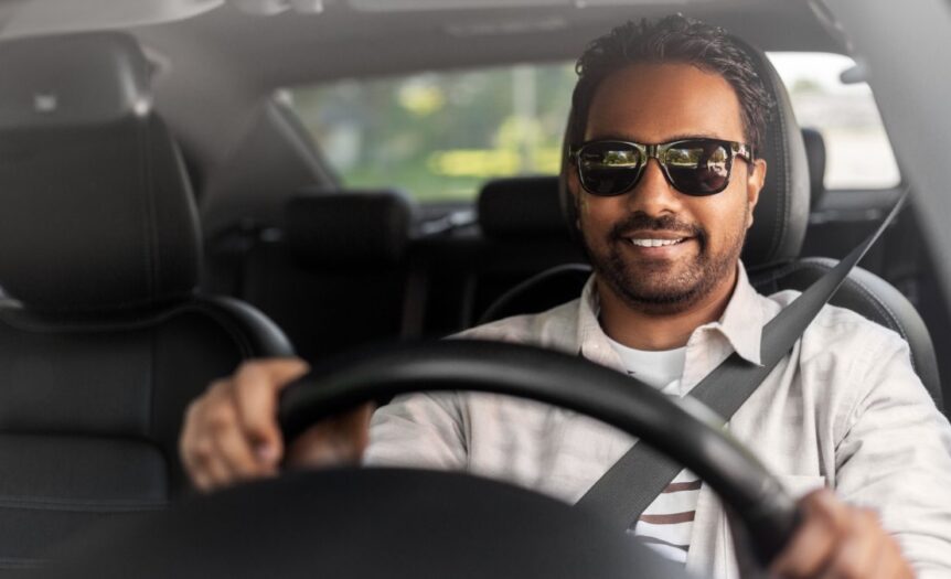 A man wearing a white shirt and black sunglasses sits in the driver's seat of a vehicle. His hands are on the steering wheel.