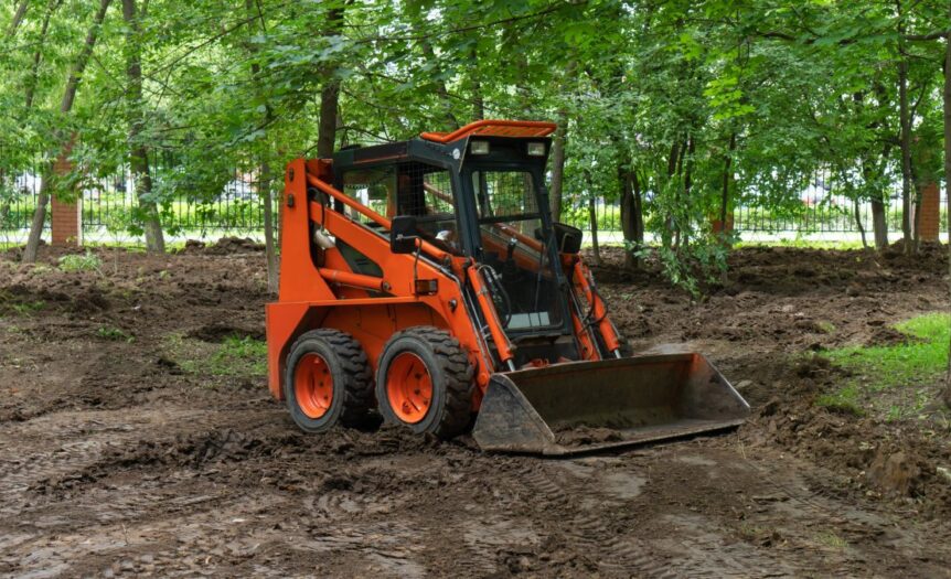 An orange skid steer loader is clearing a large site by moving soil. The site is surrounded by mature trees.