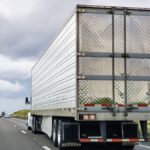 Rear view of a semi truck driving on a country road. Several cars are ahead of the truck on the road.