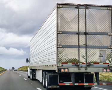 Rear view of a semi truck driving on a country road. Several cars are ahead of the truck on the road.