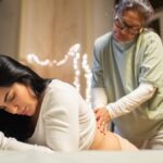 A certified nurse midwife helping a laboring mother at her bedside. There's fairy lights hanging behind them.