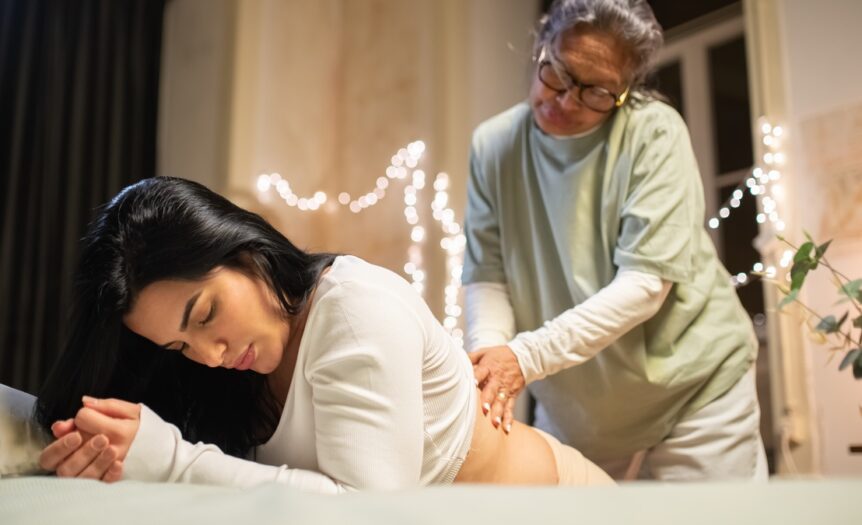 A certified nurse midwife helping a laboring mother at her bedside. There's fairy lights hanging behind them.