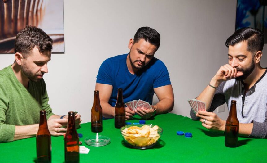 A group of men play a game of poker around a square table with green felt. On the table are playing cards and poker chips.