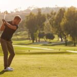 A male golfer on the green getting ready to swing down at the ball. The sun is rising behind him and he's focused.