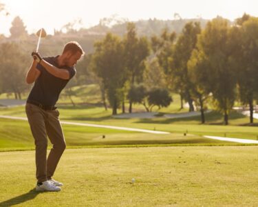 A male golfer on the green getting ready to swing down at the ball. The sun is rising behind him and he's focused.