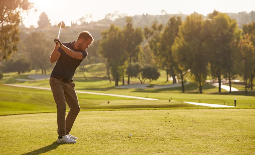 A male golfer on the green getting ready to swing down at the ball. The sun is rising behind him and he's focused.