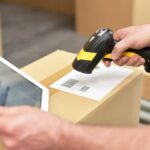 A logistics professional scanning packaging barcodes. They're wearing a yellow safety vest and holding a tablet.