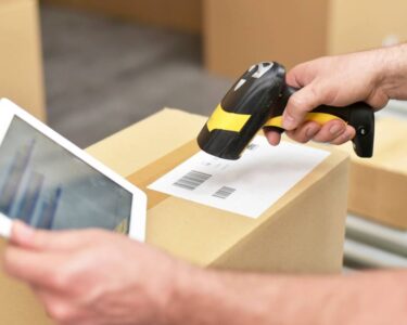 A logistics professional scanning packaging barcodes. They're wearing a yellow safety vest and holding a tablet.