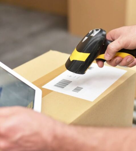 A logistics professional scanning packaging barcodes. They're wearing a yellow safety vest and holding a tablet.