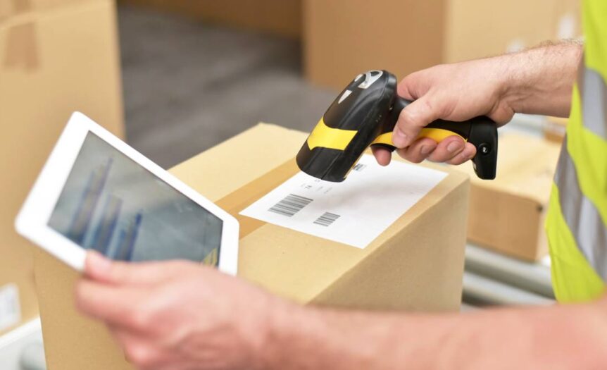 A logistics professional scanning packaging barcodes. They're wearing a yellow safety vest and holding a tablet.