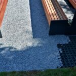 Close up on a pair of park benches set up on a patch of grey and pebbly permeable paving on a very sunny day.