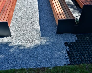 Close up on a pair of park benches set up on a patch of grey and pebbly permeable paving on a very sunny day.