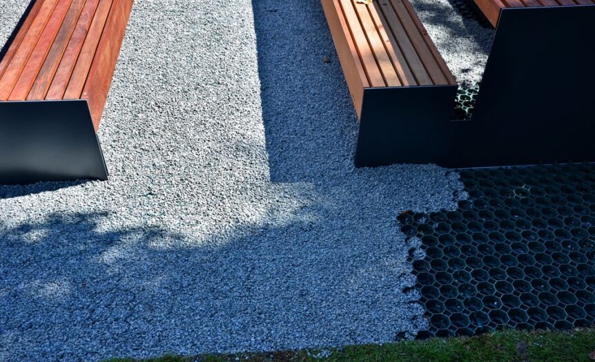 Close up on a pair of park benches set up on a patch of grey and pebbly permeable paving on a very sunny day.