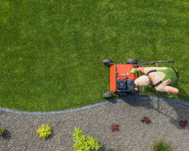 A top-down view of a landscaping professional mowing a lawn. He is mowing right up against the edge of the grass.