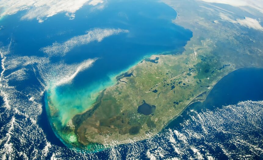 A view of the state of Florida from space. Lakes, green land, emerald-blue waters, and light cloud coverage are visible.