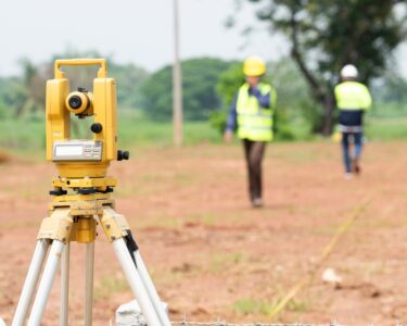 A theodolite sits on a tripod at a construction site during a land survey with workers wearing hard hats in the distance.