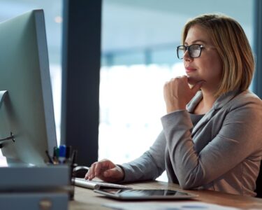 A woman sits at her desk in an office environment and reads something on her computer. She has a thoughtful look.