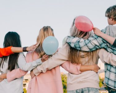 A group of seven teens are in a line with their arms around each other's shoulders; some are holding pastel balloons.