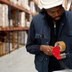 A warehouse team member wears a blue jacket and a white hard hat. They are taping a white cardboard box closed.