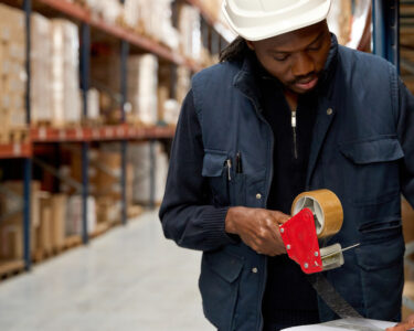 A warehouse team member wears a blue jacket and a white hard hat. They are taping a white cardboard box closed.