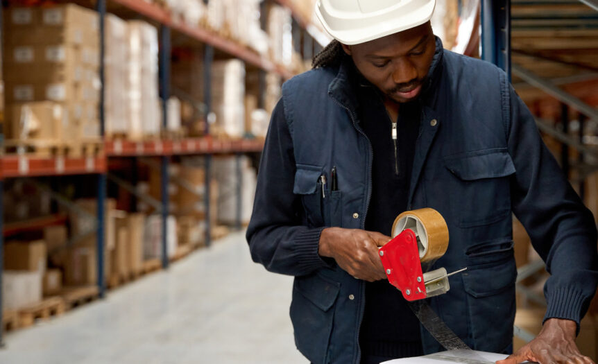 A warehouse team member wears a blue jacket and a white hard hat. They are taping a white cardboard box closed.