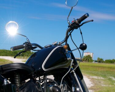 A black motorcycle is on a dirt road in a grassy field near a resort building. The sun shines through a motorcycle mirror.
