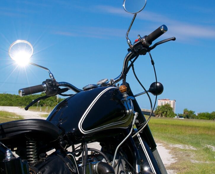 A black motorcycle is on a dirt road in a grassy field near a resort building. The sun shines through a motorcycle mirror.