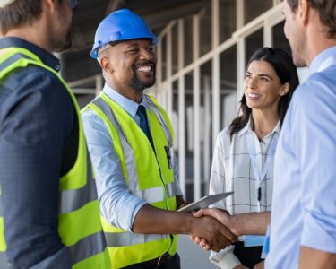 A person in a high-vis safety vest and a blue hard hat shakes hands with a man in a dress shirt. Two other people stand by.