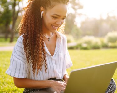 A woman using airpods in professional attire works on her computer, sitting on the grass on a sunny day.