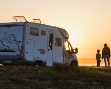 An adult and child hold hands while they watch the sunset over a body of water from next to their RV.