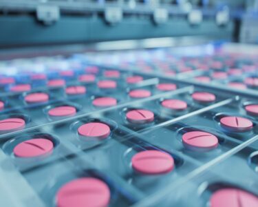 A close up of pink, circular pills being packed into plastic packaging in an assembly line.