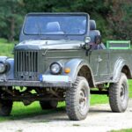 A vintage World War II–era military jeep parked in the middle of a gravel road. The jeep has a dark green paint color.