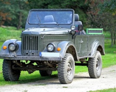 A vintage World War II–era military jeep parked in the middle of a gravel road. The jeep has a dark green paint color.