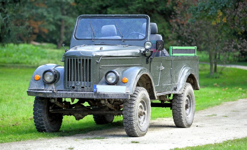 A vintage World War II–era military jeep parked in the middle of a gravel road. The jeep has a dark green paint color.