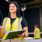 A woman standing in front of an outdoor stage wearing a safety vest and headset while holding a two-way radio and clipboard.