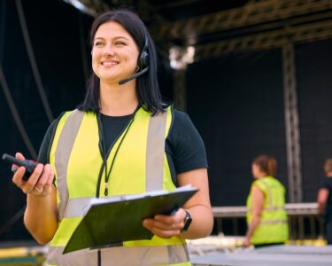 A woman standing in front of an outdoor stage wearing a safety vest and headset while holding a two-way radio and clipboard.
