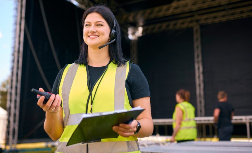A woman standing in front of an outdoor stage wearing a safety vest and headset while holding a two-way radio and clipboard.