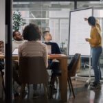 Four people sit at a table in a conference room, watching a woman standing and pointing at a whiteboard.