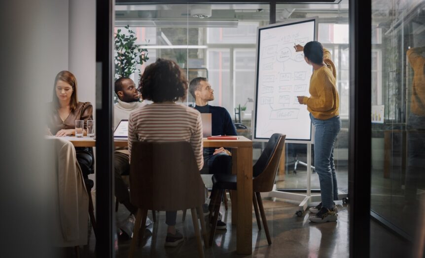 Four people sit at a table in a conference room, watching a woman standing and pointing at a whiteboard.