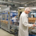 A man wearing a white lab coat, a hair net, and safety goggles looks at a large touchscreen on a packaging machine.