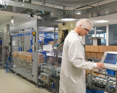 A man wearing a white lab coat, a hair net, and safety goggles looks at a large touchscreen on a packaging machine.