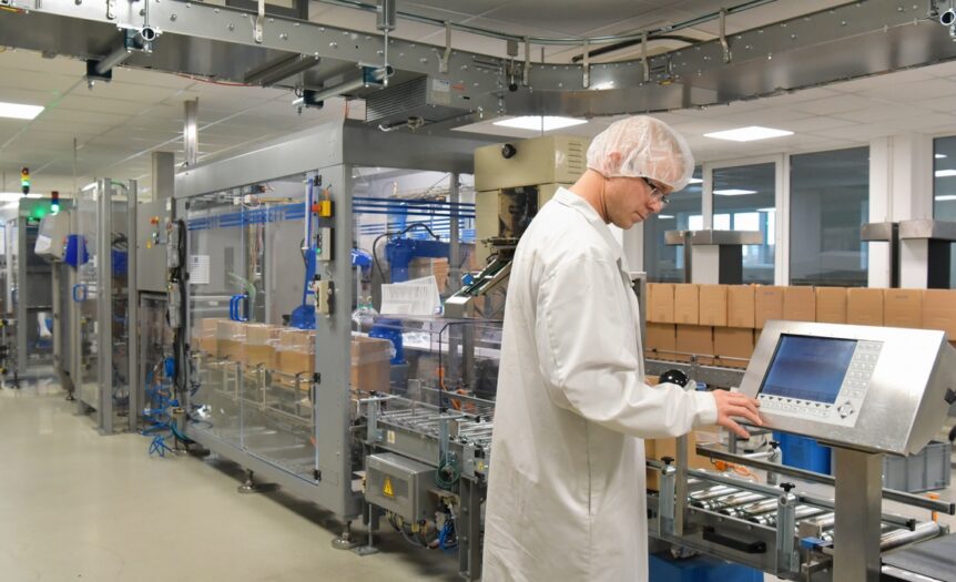 A man wearing a white lab coat, a hair net, and safety goggles looks at a large touchscreen on a packaging machine.