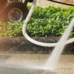 A close-up view shows a person wearing brown pants and black boots pressure washing a concrete surface.