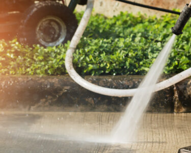 A close-up view shows a person wearing brown pants and black boots pressure washing a concrete surface.