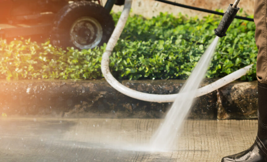 A close-up view shows a person wearing brown pants and black boots pressure washing a concrete surface.
